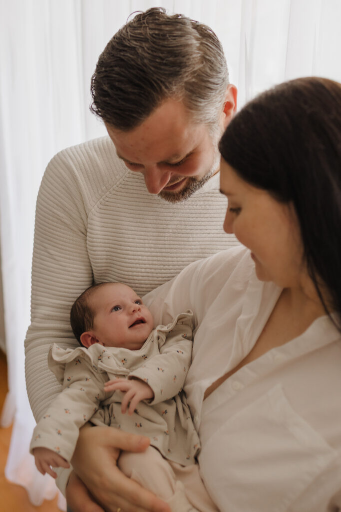 Neugeborenenshooting in Lübeck. Mama und Papa halten das Baby im Arm. Das Baby lächelt die Eltern an.