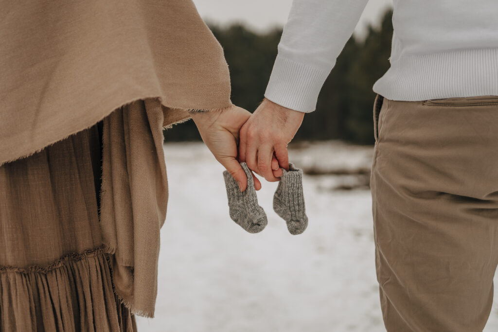 Wintershooting in der Palinger Heide. Babybauchshooting. Die Eltern gehen Hand in Hand spazieren und halten Babysocken in den Händen.